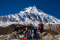 Group of trekkers posing on rocky terrain below the majestic snow-covered Manaslu peak near Larke Pass, with the summit rising clearly against a deep blue Himalayan sky.