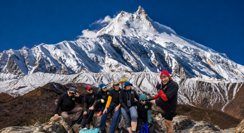 Group of trekkers posing on rocky terrain below the majestic snow-covered Manaslu peak near Larke Pass, with the summit rising clearly against a deep blue Himalayan sky.